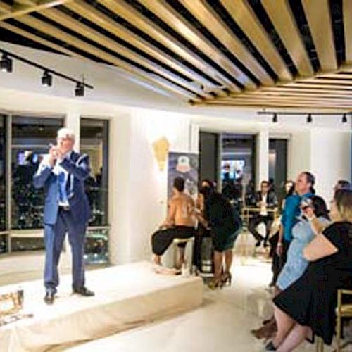 A person in a suit is speaking on a platform in front of an engaged audience in a modern, well-lit room with large windows and wooden ceiling beams.