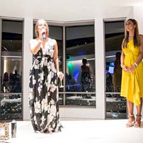 Two women are standing in a well-lit room with large windows at an event. The woman in the floral dress is speaking into a microphone, and the other in yellow looks on.