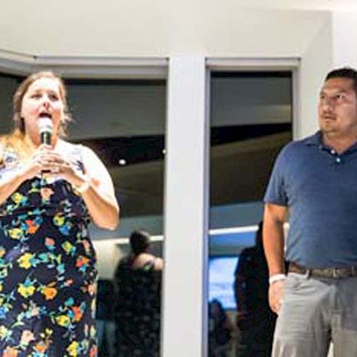 A woman holds a microphone while speaking, and a man stands beside her, both in casual clothing in an indoor setting with people in the background.