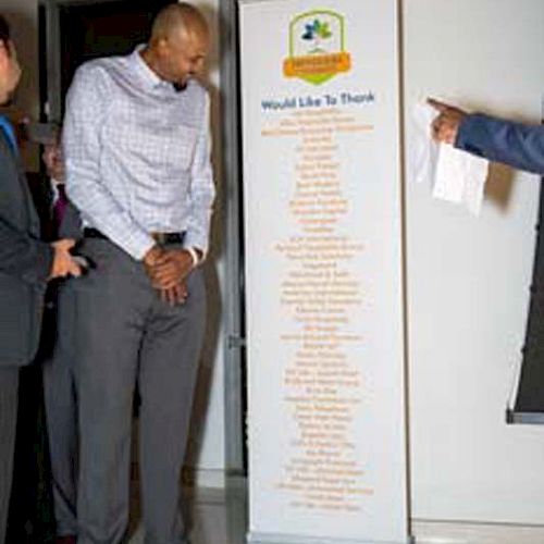 A tall man standing next to a banner with text, while another person points at the banner. The setting appears to be indoors.