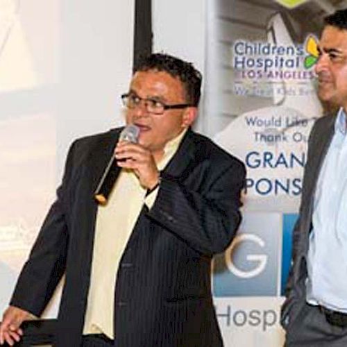 Two men are standing, one holding a microphone, with a backdrop that includes a banner for Children's Hospital Los Angeles.