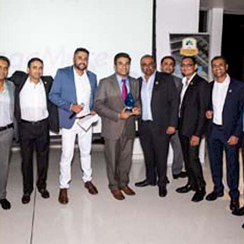 A group of men, some dressed in suits, pose together indoors, with one man holding a trophy, possibly at an event or awards ceremony.