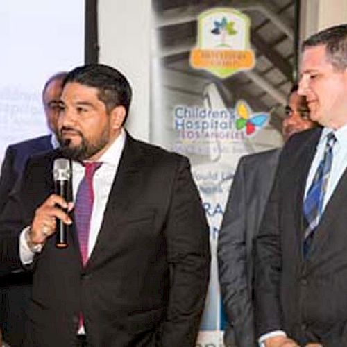 A man in a suit is speaking into a microphone while others in suits stand nearby at an event related to Children’s Hospital of Los Angeles.