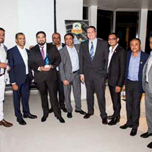 A group of men dressed in formal attire are standing together, with one holding a trophy. They are posing for a group photo indoors, smiling.