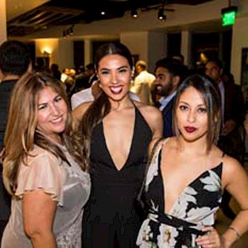 Three women are posing for a photo at a social event. The background shows other attendees mingling and some event signage.