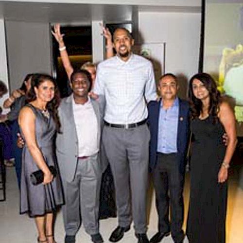 A group of five people dressed formally, posing for a photo at an event. A woman in the background has raised arms.