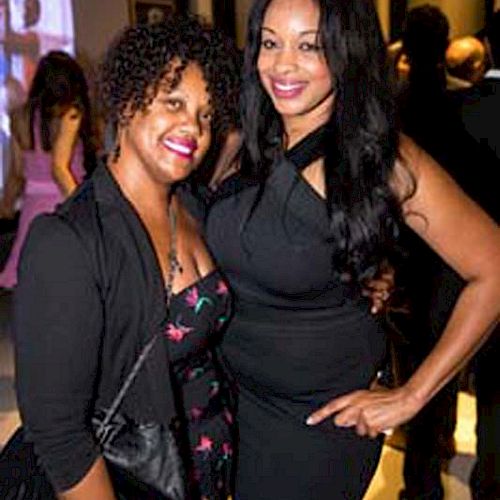 Two women are posing together at an indoor event. One is wearing a black outfit with a floral pattern, and the other is in a sleeveless black dress.