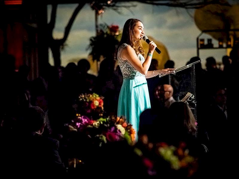 A woman in a light blue dress is speaking into a microphone at an event, with floral arrangements visible on the tables around her.