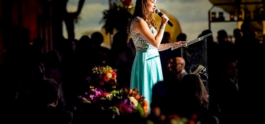A woman in a light blue dress is speaking into a microphone at an event, with floral arrangements visible on the tables around her.