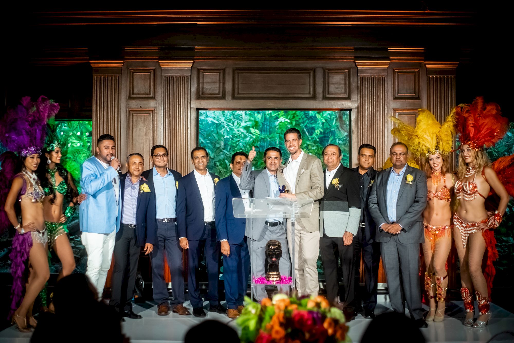 A group of people, including men in suits and women in colorful costumes, stand on a stage with a decorative backdrop.