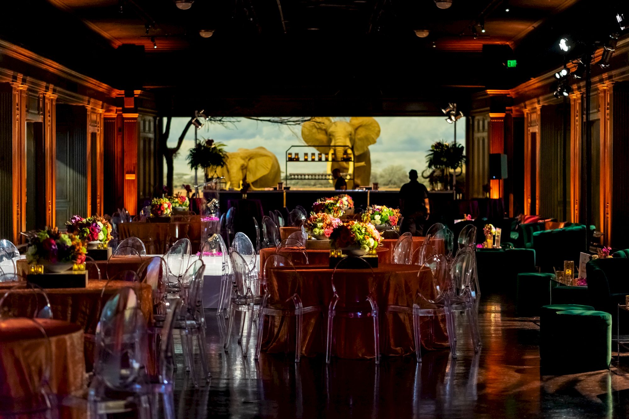 The image shows a sophisticated, dimly lit event space with decorated round tables, floral centerpieces, clear chairs, and a backdrop of elephant statues.