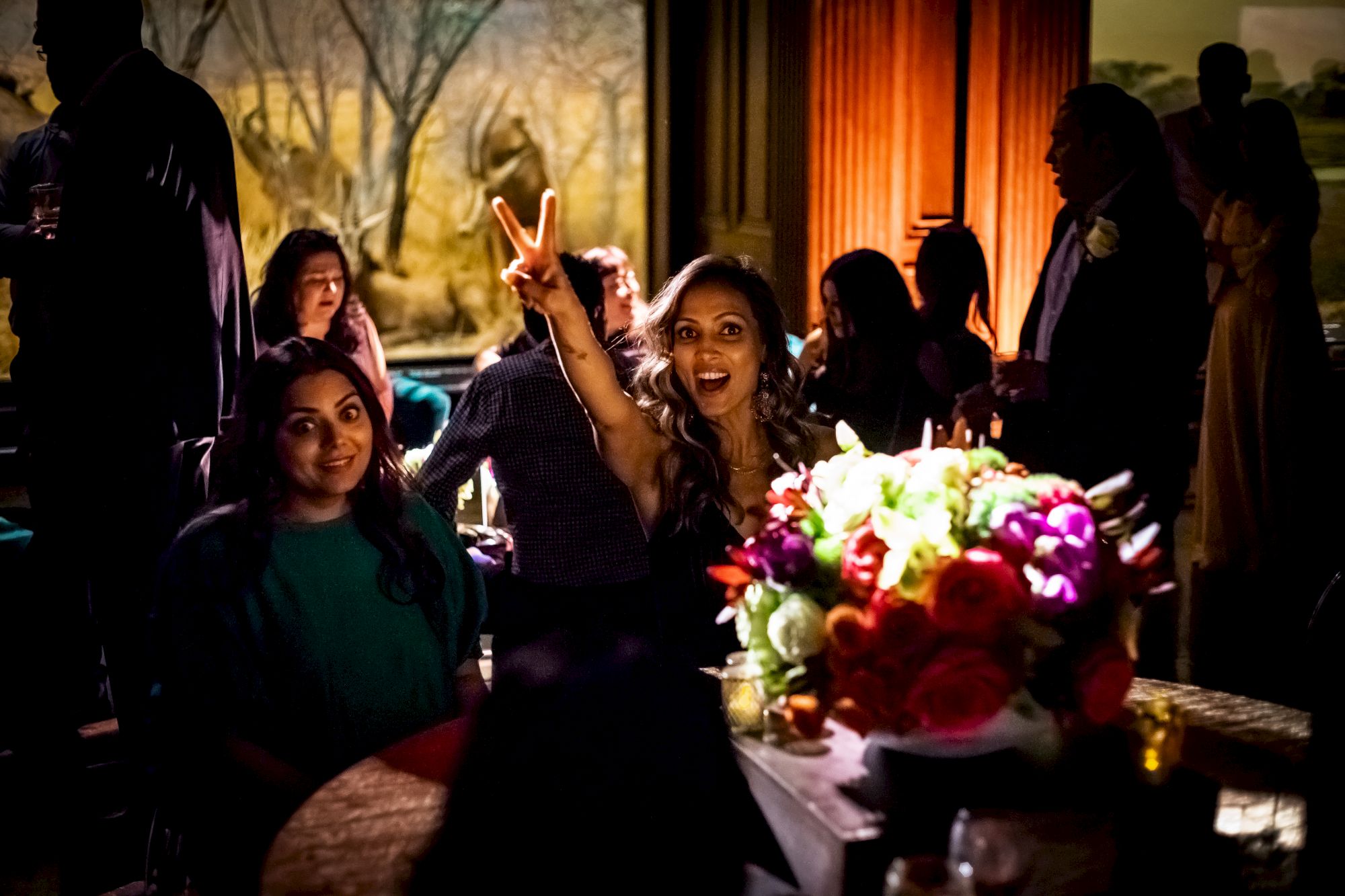 A group of people are socializing in a dimly lit room; one woman in the center is enthusiastically making a peace sign with her fingers.