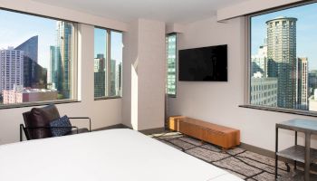 This image shows a modern hotel room with large windows, a wall-mounted TV, a bed, and an armchair, overlooking a cityscape in the background.