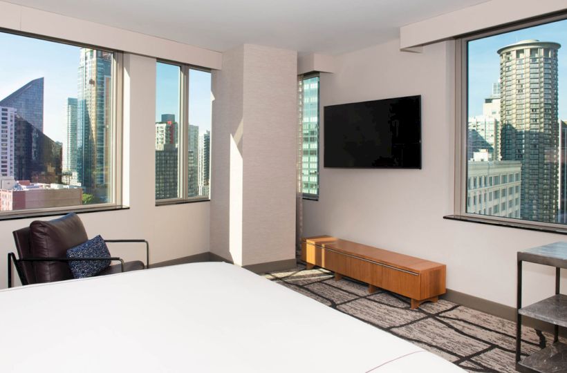 This image shows a modern hotel room with large windows, a wall-mounted TV, a bed, and an armchair, overlooking a cityscape in the background.
