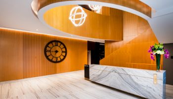 A modern reception area with a marble desk, wood paneling, a large wall clock, a staircase, and decorative light fixtures and flowers.