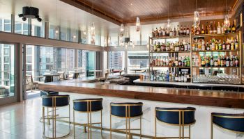 The image shows a modern bar with a well-stocked liquor shelf, stylish seating at the counter, and large windows offering a city view.