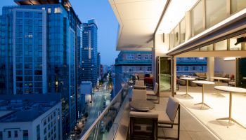 The image shows a modern urban balcony overlooking a cityscape with tall buildings at dusk. There are tables and chairs on the balcony.