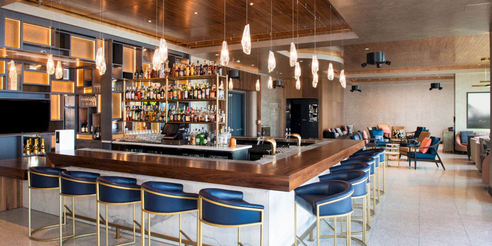The image shows a modern bar with a wooden counter, blue cushioned bar stools, and a variety of liquor bottles on shelves behind the bar.