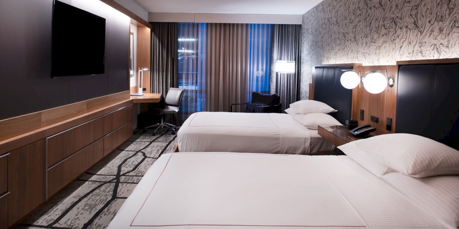 A modern hotel room with two double beds, a large flat-screen TV, a desk, and contemporary decor. The room has a monochromatic theme and large windows.