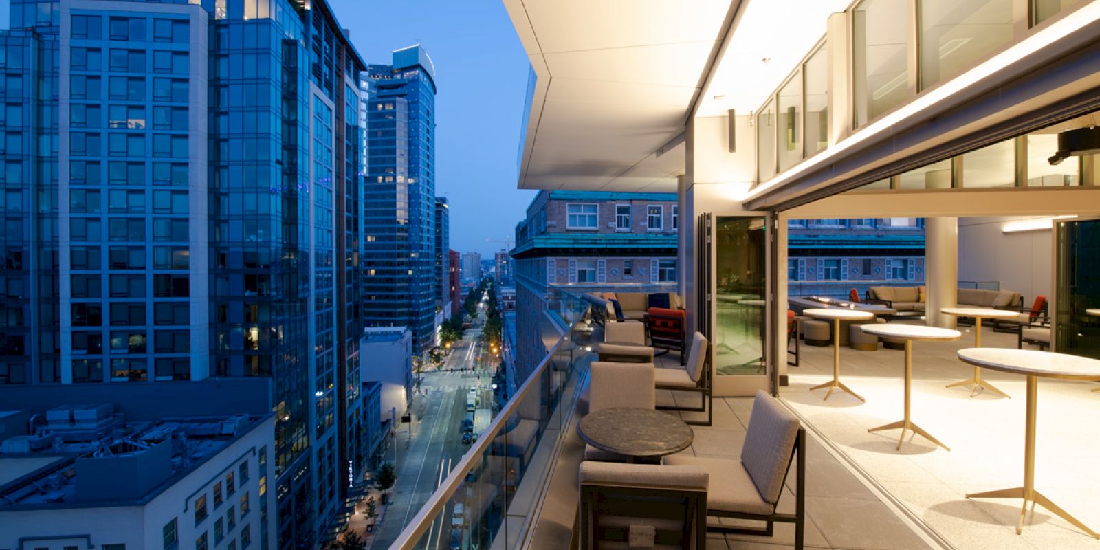 A modern balcony overlooking a city street with high-rise buildings at dusk. The balcony features seating areas and tables.