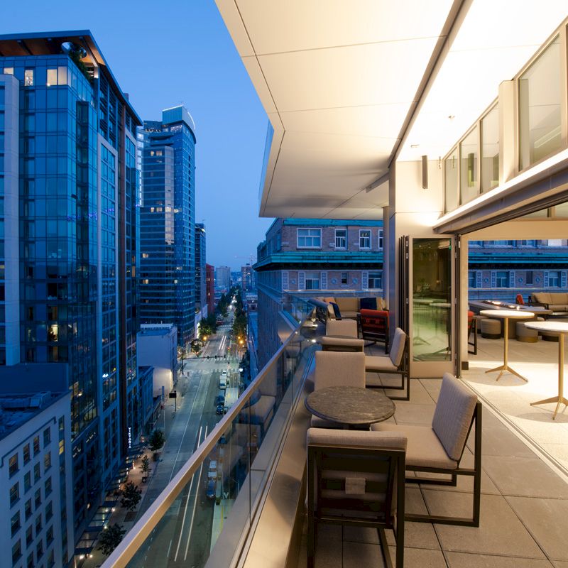 The image shows a modern rooftop deck with seating areas overlooking a cityscape with tall buildings at dusk or evening.