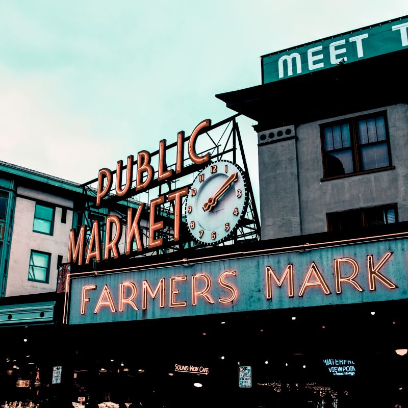 The image shows an outdoor scene with neon signs for "PUBLIC MARKET" and "FARMERS MARKET" on buildings, featuring a clock and various shops below.