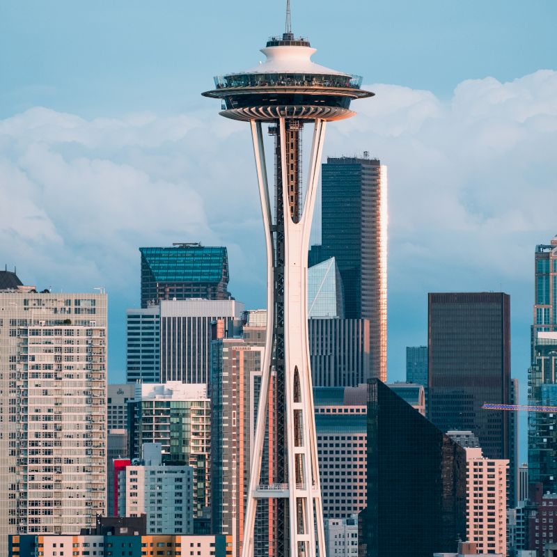 The image shows the Space Needle with a backdrop of downtown Seattle, featuring various modern skyscrapers and buildings under a partly cloudy sky.