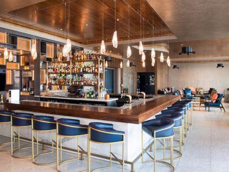 This image shows a modern bar with blue barstools, pendant lights, and a well-stocked back bar, located within an open, well-lit lounge area.
