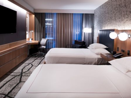 The image shows a modern hotel room with two beds, a flat-screen TV, a work desk, and stylish lighting and decor, creating a cozy atmosphere.