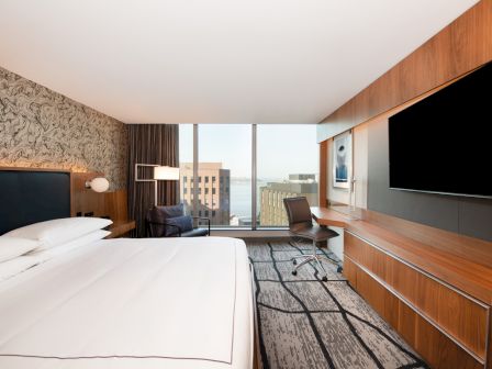 The image shows a modern hotel room with a large bed, a wall-mounted TV, a desk, and a window overlooking a cityscape with buildings and water.