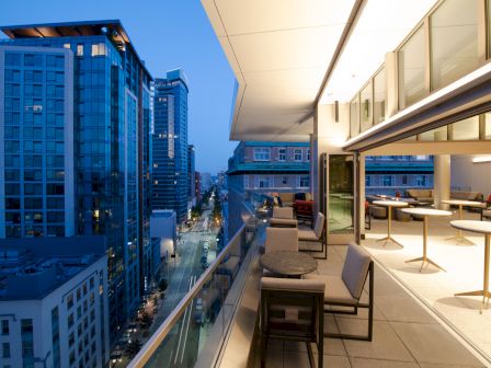 A modern cityscape during twilight with tall buildings, viewed from a high balcony of an upscale restaurant or lounge with tables and chairs.