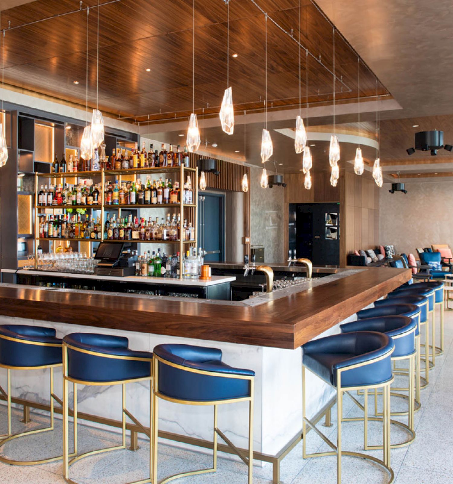 This image shows a modern bar with high chairs, a stocked bar, pendant lights, and seating areas in a stylish, contemporary setting.