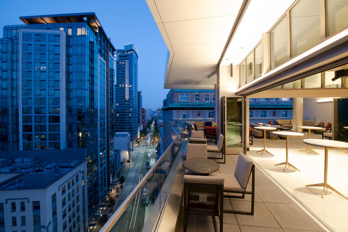 The image shows a modern cityscape during the evening, with high-rise buildings and a well-lit balcony featuring outdoor seating and tables.