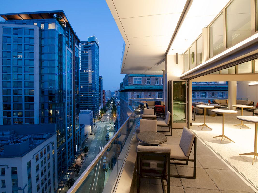 The image shows a modern cityscape during the evening, with high-rise buildings and a well-lit balcony featuring outdoor seating and tables.