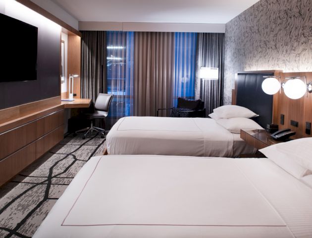 The image shows a modern hotel room with two neatly made beds, a large TV, a desk with a chair, and lamps, featuring contemporary decor and design.