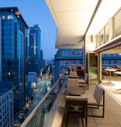 A modern balcony view at dusk showing city skyscrapers, a stylish seating area, and large windows revealing the indoor space.