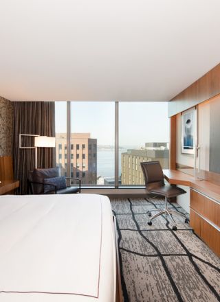 A modern hotel room features a king-sized bed, large TV, desk, chair, artwork, and a window with a city view in the background.
