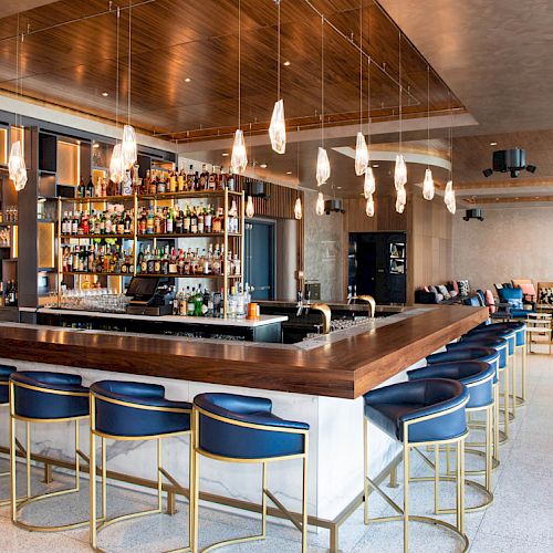 A modern bar with blue barstools, a well-stocked back shelf, elegant hanging lights, and a seating area in the background.