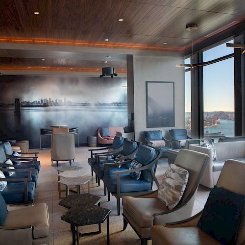 This image shows a modern lounge with comfortable seating, large windows with a scenic view of water and a Ferris wheel, and contemporary decor.
