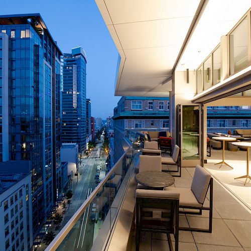 A modern balcony overlooking a city street at dusk, with seating and tables arranged. Nearby are tall, illuminated buildings.