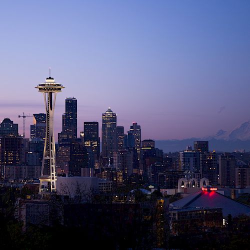 A cityscape at dusk featuring the Space Needle, downtown buildings, and a distant mountain under a twilight sky.