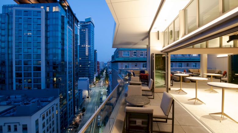 A modern cityscape at dusk, featuring high-rise buildings adjacent to a sleek, open-air balcony with seating and tables, illuminated by warm lights.