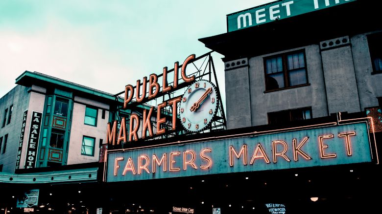 This image shows the iconic neon signs for the Public Market and Farmers Market at Pike Place Market in Seattle, with adjacent buildings in the background.