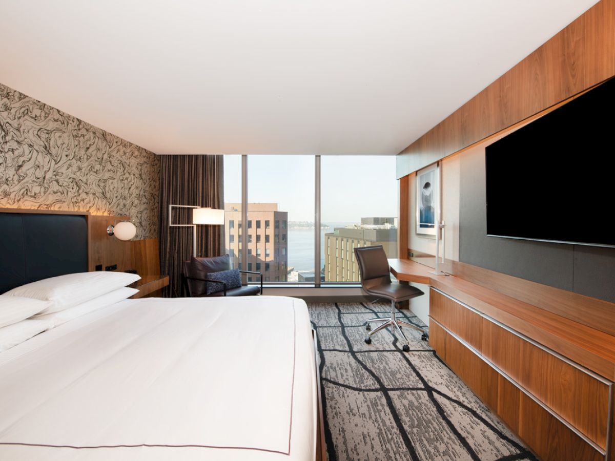 A modern hotel room with a large bed, wall-mounted TV, desk, chair, and a window offering a city and water view.