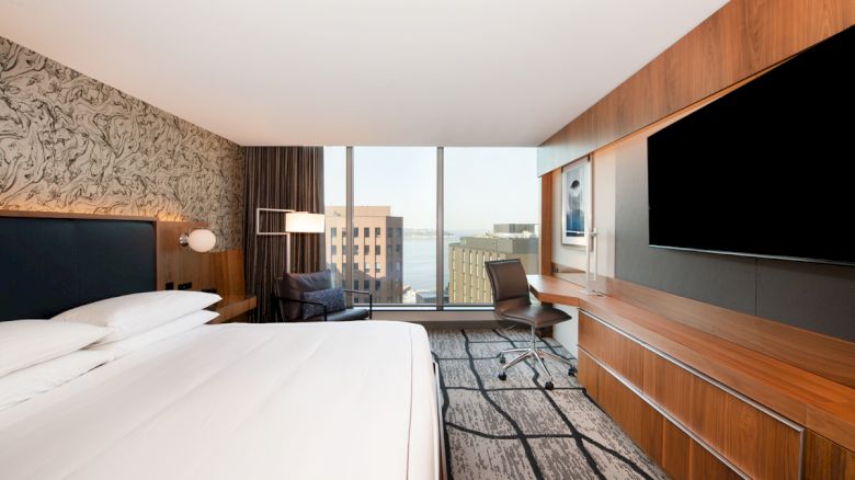 A modern hotel room with a large bed, wall-mounted TV, desk, chair, and a window offering a city and water view.