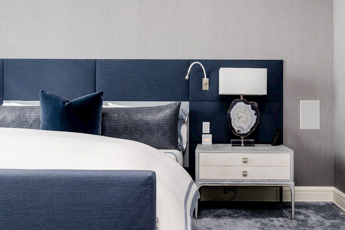 A modern bedroom with a blue and white color scheme, featuring a bed with blue upholstery and a nightstand with a lamp and decorative items.