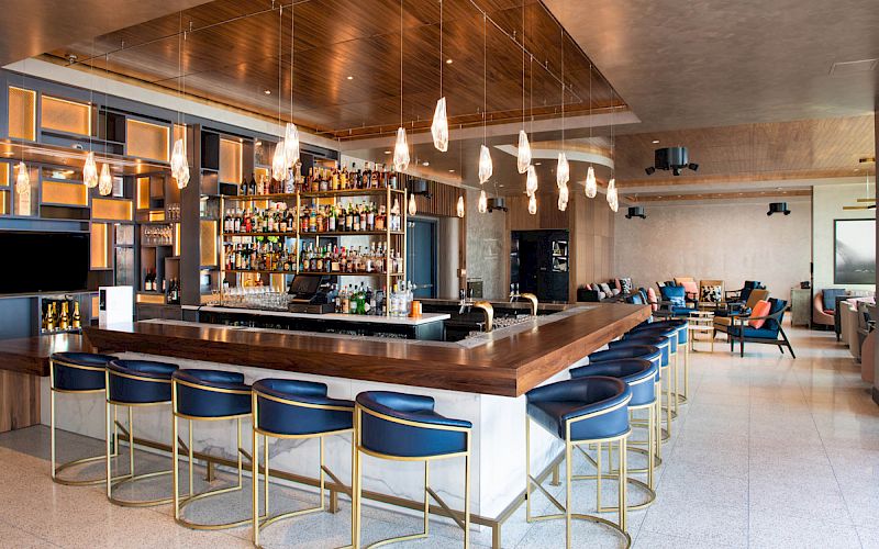 The image shows a modern bar with a well-stocked liquor display, blue and gold barstools, pendant lights, and a seating area in the background.