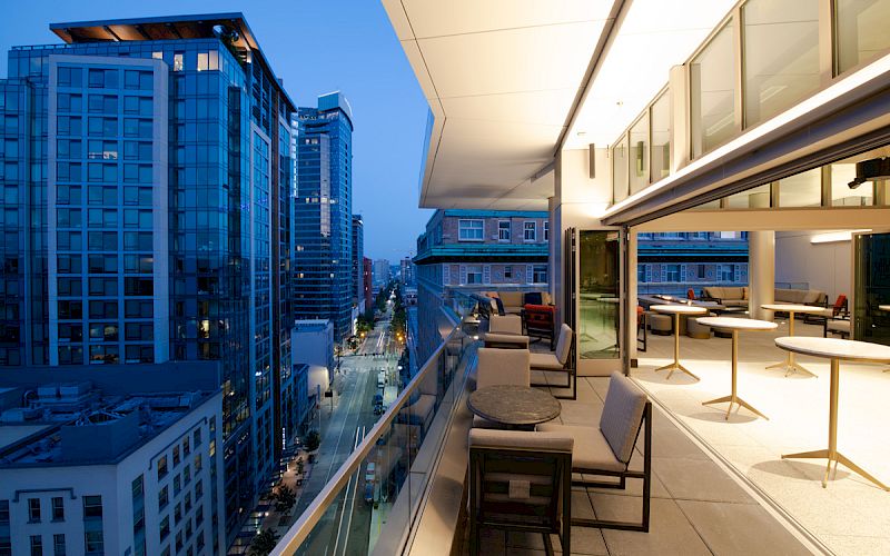 A modern cityscape with tall buildings is viewed from a balcony in the evening, featuring outdoor seating and tables with a spacious design.