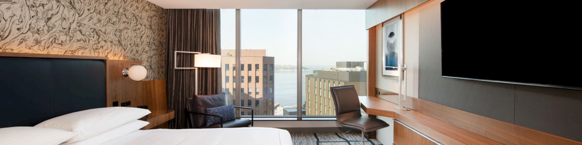A modern hotel room with a large bed, a wall-mounted TV, a desk with a chair, and a window overlooking buildings and a body of water.