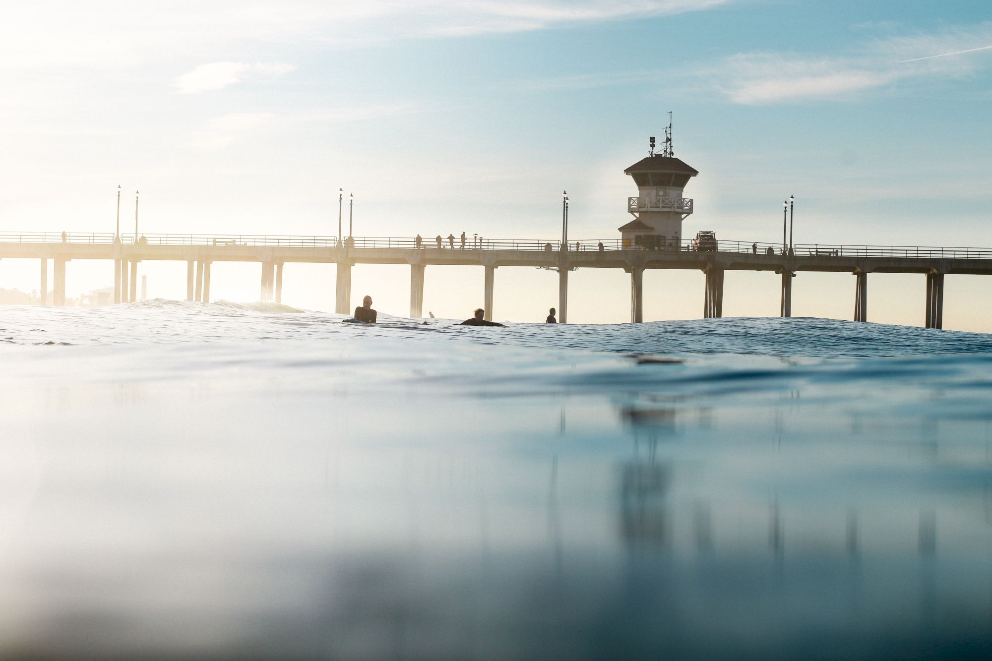 The image shows a calm ocean scene with surfers in the water and a pier in the background stretching into the horizon.
